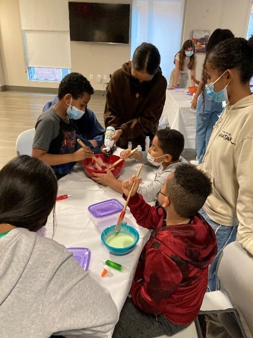 Children wearing masks, seated on a table, are preparing a kind of colored gum.