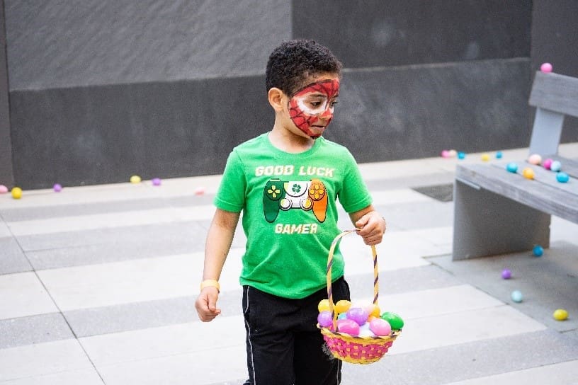 Boy dressed in a green polo shirt, carrying a basket with eggs in his hand, his face is painted spider man.
