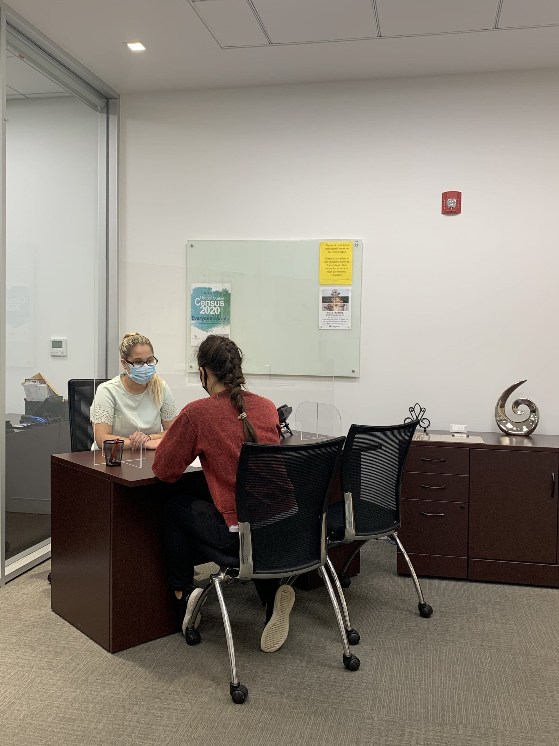 Two women conversing, wearing masks, seated leaning against a desk.